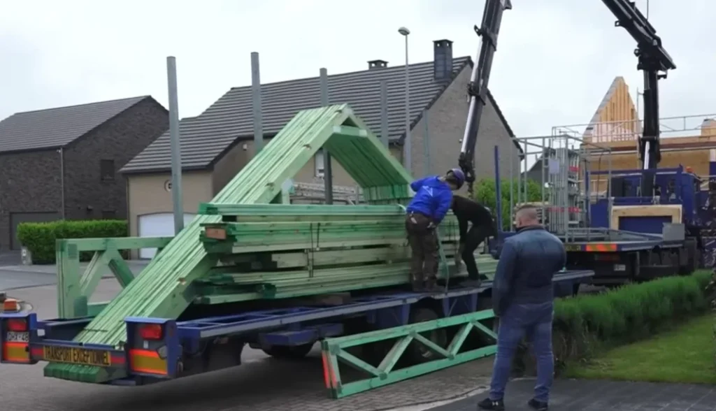 Um caminhão com guindaste descarregando paletes com grandes vigas de madeira triangulares pré-cortadas e verdes para a montagem do telhado de uma casa Gablok.