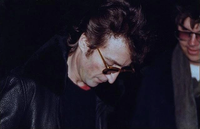 Foto histórica de John Lennon de perfil, usando óculos, assinando o álbum Double Fantasy para o seu assassino Mark Chapman, que aparece parcialmente ao fundo.