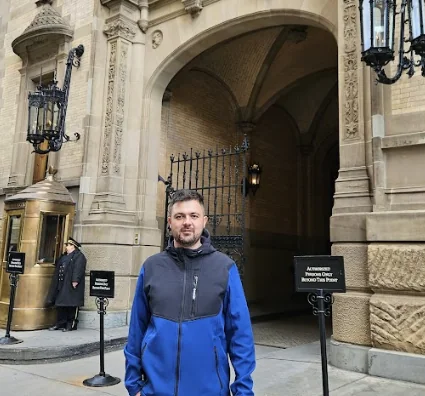 Alexandre Cordeiro, fundador do Inacreditável Mundo Curioso, vestindo uma jaqueta azul e preta, em frente ao arco de entrada de pedra do Edifício Dakota em Nova York.
