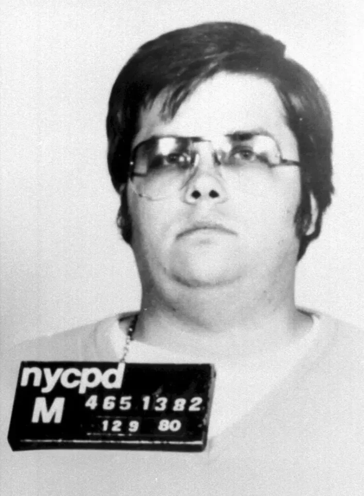 Foto de fichamento policial em preto e branco de Mark David Chapman jovem, usando óculos, após ser preso pelo assassinato de John Lennon em Nova York.