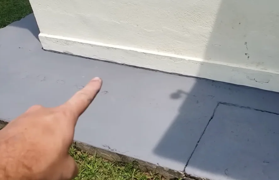 Close em uma calçada pintada de cinza claro; uma mão aparece no canto esquerdo da imagem apontando o dedo indicador para o detalhe de um mini rodapé, onde a tinta de piso sobe discretamente cerca de dois centímetros na base de um muro branco lateral para esconder imperfeições.