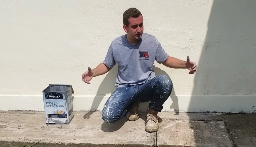 Um homem agachado em uma calçada de cimento ao ar livre, apontando com os polegares para uma lata de tinta para pisos e cimentados da marca Leinertex posicionado ao seu lado esquerdo, rente a um muro branco ao fundo.