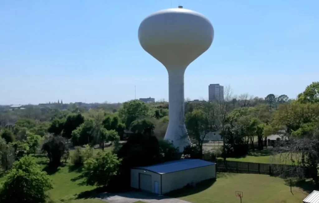 Grande torre de água pública (caixa d'água urbana) em formato de cálice pintada de branco, erguida acima de uma área arborizada com árvores verdes sob um céu azul claro nos Estados Unidos. (Foto: Canal RicardoMolinaUSA)