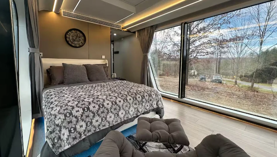 Vista interna do quarto da mini casa cápsula inteligente, mostrando uma cama de casal arrumada com colcha estampada em tons de cinza e preto, ao lado de uma imensa parede de vidro panorâmica que revela as árvores do bosque na parte externa.