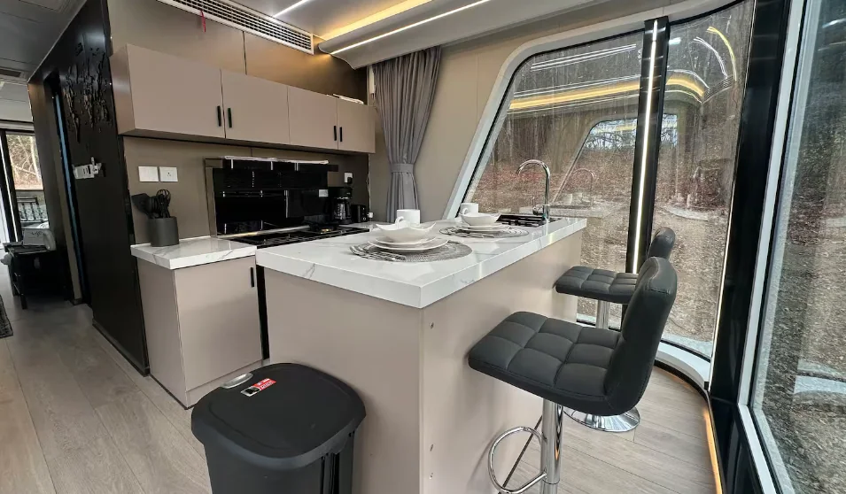 Vista interna da cozinha moderna e minimalista da mini casa cápsula, com destaque para uma ilha central branca com duas banquetas pretas de couro, armários planejados ao fundo com iluminação LED embutida e uma grande janela de vidro com vista para o exterior.