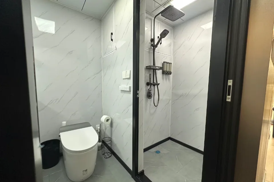 Vista interna do banheiro inteligente da mini casa cápsula, com paredes revestidas em placas de plástico brancas imitando mármore, vaso sanitário smart tecnológico à esquerda e área do box aberta ao fundo com um chuveiro de teto em estilo chuva.