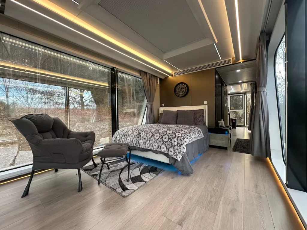 Perspectiva ampla do interior do quarto principal da mini casa cápsula inteligente, mostrando o piso laminado de madeira, uma poltrona cinza à esquerda, a cama de casal ao fundo e o teto moderno com fitas de iluminação LED embutidas lineares e amareladas.