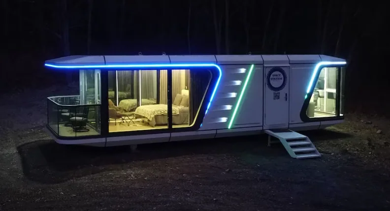 Fotografia noturna da mini casa cápsula inteligente totalmente iluminada, exibindo linhas de LED em neon azul ao redor da estrutura externa cinza e grandes painéis de vidro acesos com luz quente no interior.