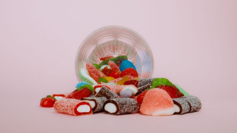 Diversos doces, guloseimas coloridas e balas de goma espalhadas em frente a uma tigela de vidro em fundo rosa.
